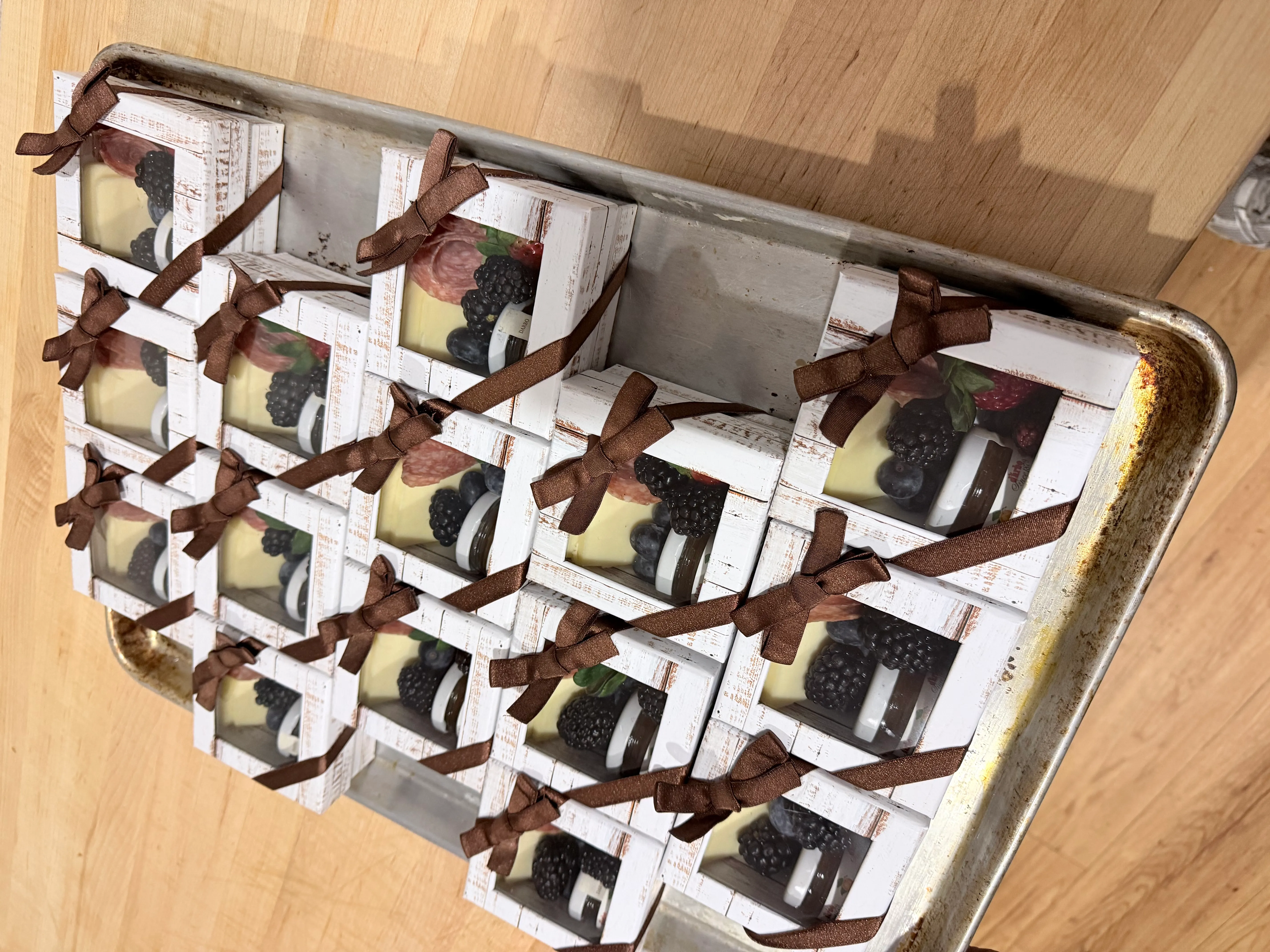 A tray of individually packaged rustic white charcuterie gift boxes tied with brown satin ribbons, containing fresh blackberries, blueberries, cured salami, gourmet cheese, and honey jars for VIP corporate drop-off catering.