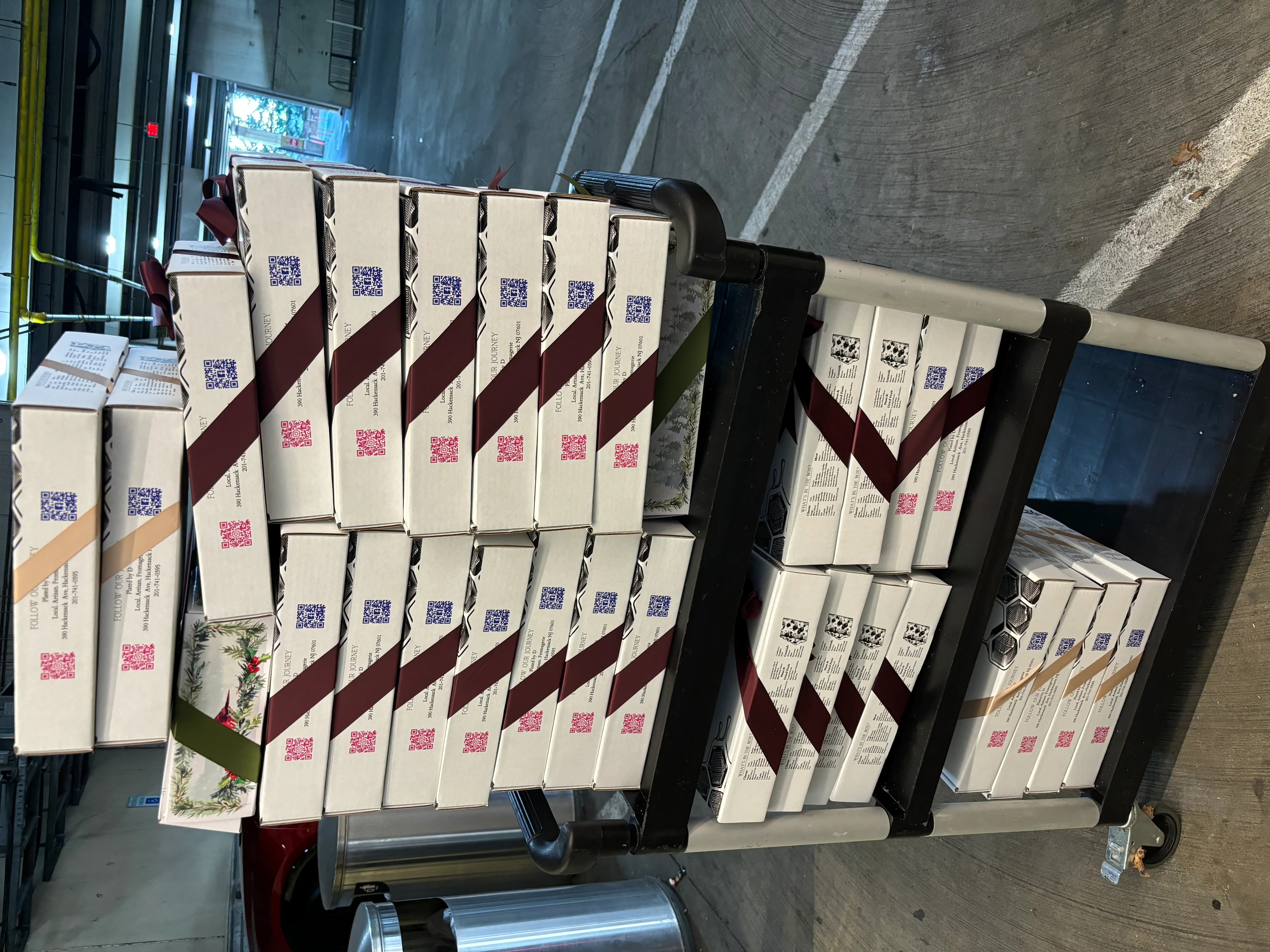 A black multi-tiered rolling cart fully loaded with elegant individually packaged corporate catering boxes tied with burgundy and green ribbons, ready for executive drop-off delivery.